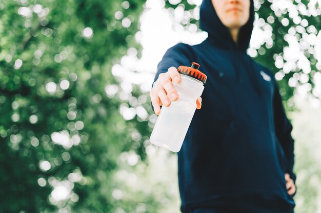 Photo close up of young man runner in hoodie holding water bottle while doing workout in summer sunny