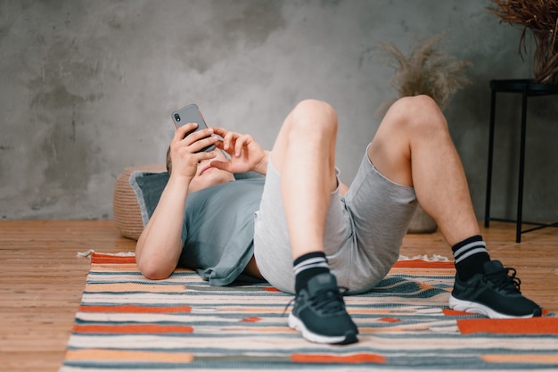 Крупным планом - молодой человек в спортивной форме отдыхает на полу дома и смотрит в телефон в социальной сети. Студент бездельничает и откладывает работу и учебу