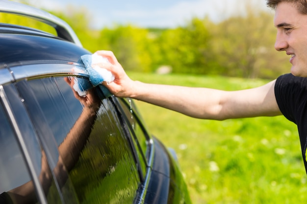 Close Up of Young Man Washing Windows of Black Luxury Vehicle with Soapy Sponge in Green Field on Bright Sunny Day