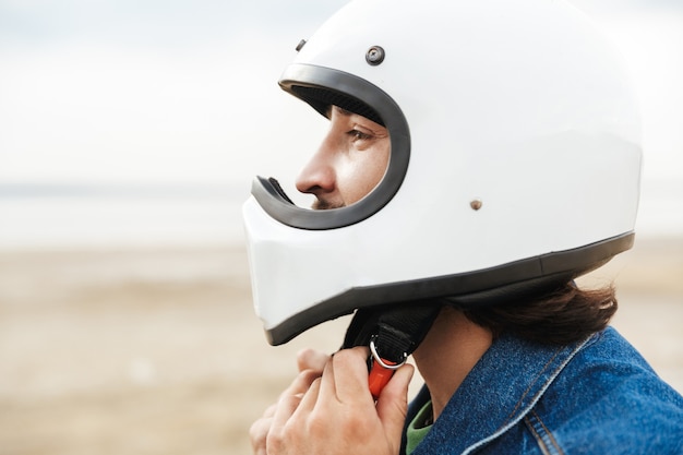 Close up of a young man wearing casual outfit at the beach, putting on a helmet