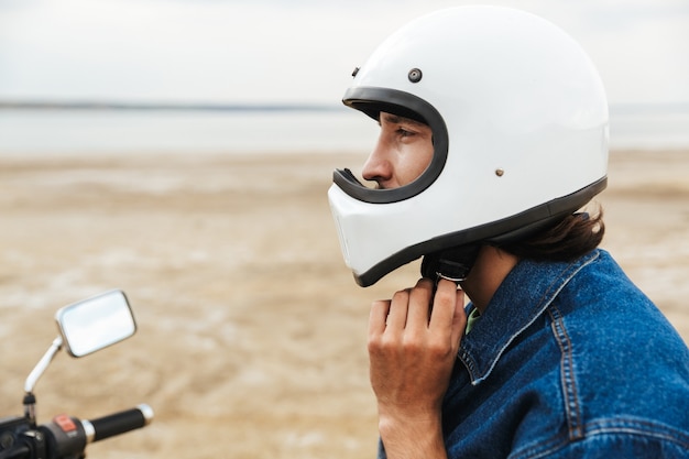 Close up of a young man wearing casual outfit sitting on a motocycle at the beach, putting on a helmet