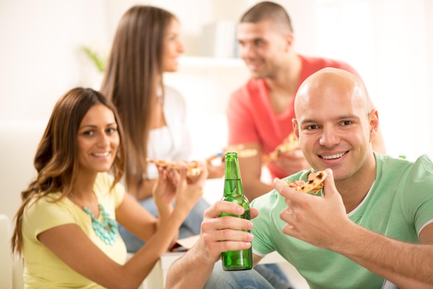 Close up of a young men smiling and eating pizza and drinking bear with her friends in the background.