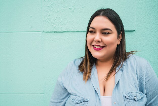 Close-up of young plus size woman smiling while standing against light blue wall outdoors.