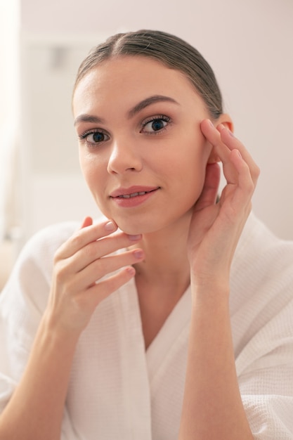 Close up of young pretty lady standing alone in white bathrobe and touching the skin on her face while smiling