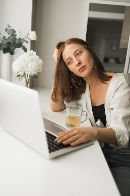 Primo piano di una giovane donna graziosa che beve tè durante la pausa dopo aver digitato e navigato online sul laptop w