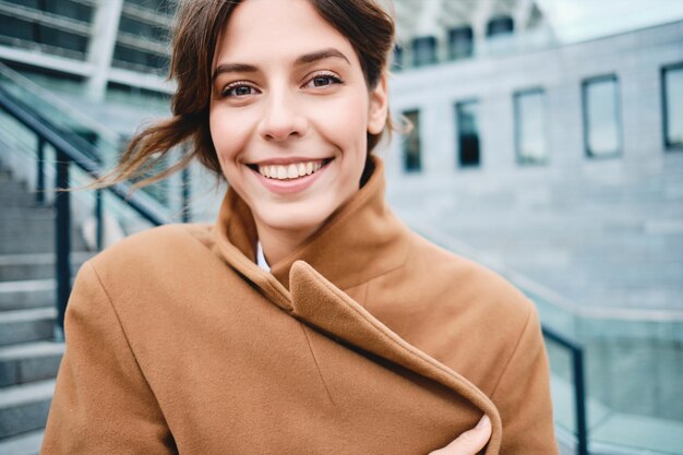 屋外のカメラで楽しく見て寒さからコートを締める若いきれいな女性をクローズアップ