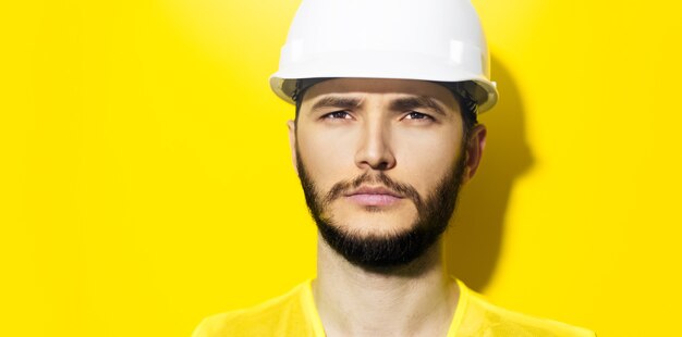 close-up young serious man architect, builder engineer, wearing white construction safety helmet and yellow jacket isolated on yellow wall.