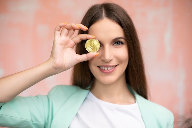 Close up young smiling female is holding cryptocurrency in front eye. Online virtual future currency