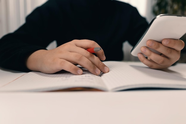 Close up of young student with smartphone  writing to notebook.