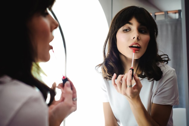 Foto close-up di una giovane donna che applica rossetto rosso davanti allo specchio a casa