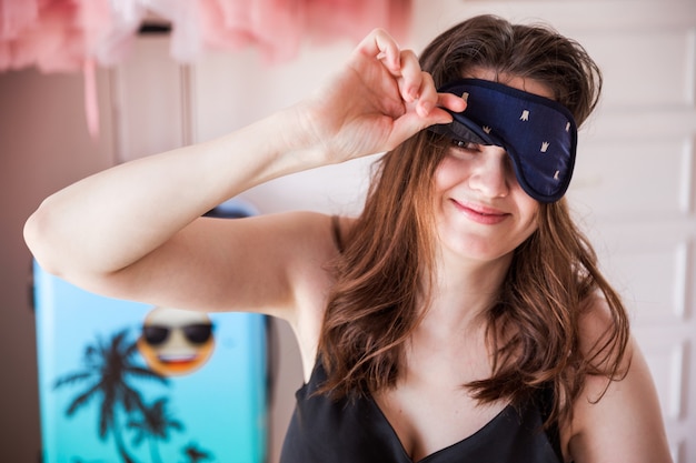 Close-up of young woman in bedroom in morning with sleeping mask , looking at camera with positive smile, enjoying free time in bedroom.  