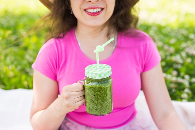 若い女性のクローズアップは、公園で楽しんだり、ピクニックで緑のスムージーを飲みます。