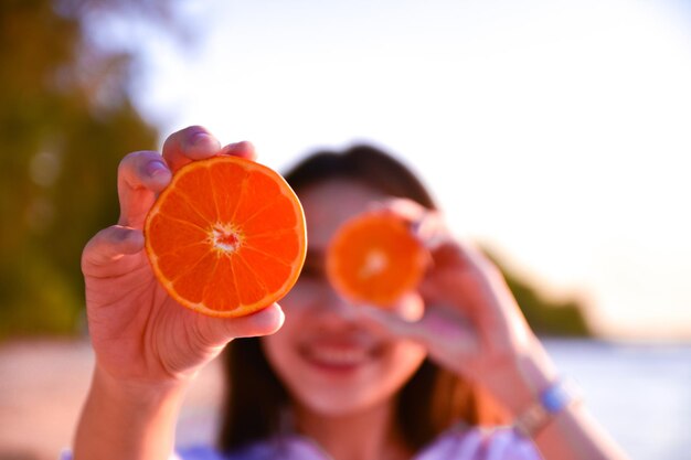 Photo close-up of young woman holding orange slices at beach against clear sky during sunset