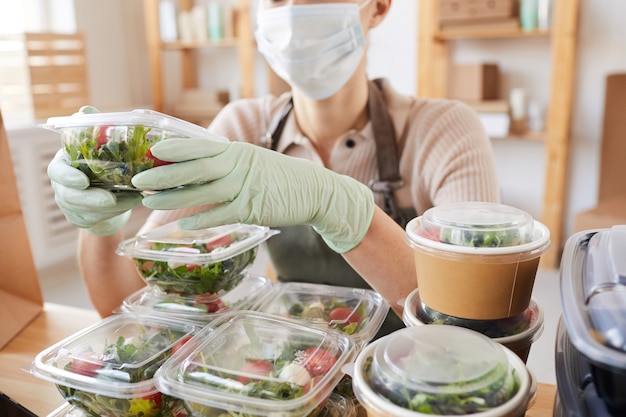 Close-up of young woman in protective mask and gloves packing fresh vegetables in boxes while sitting at the table