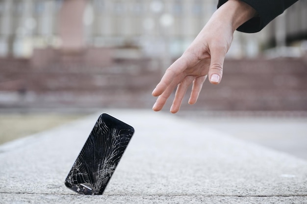 Крупным планом молодая женщина тянется к своему сломанному смартфону с треснувшим стеклом экрана