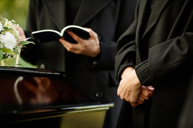 Close up of young woman standing by coffin at outdoor funeral ceremony priest reading prayer in back