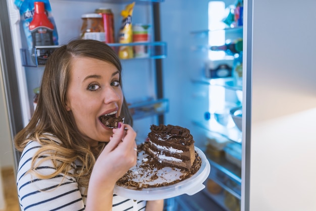 Close-up of young woman standing in front of open refrigerator eating cake 