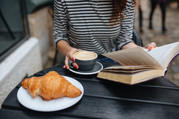 Close up of young woman in striped sweater reading book and drinking coffee at cafe terrace