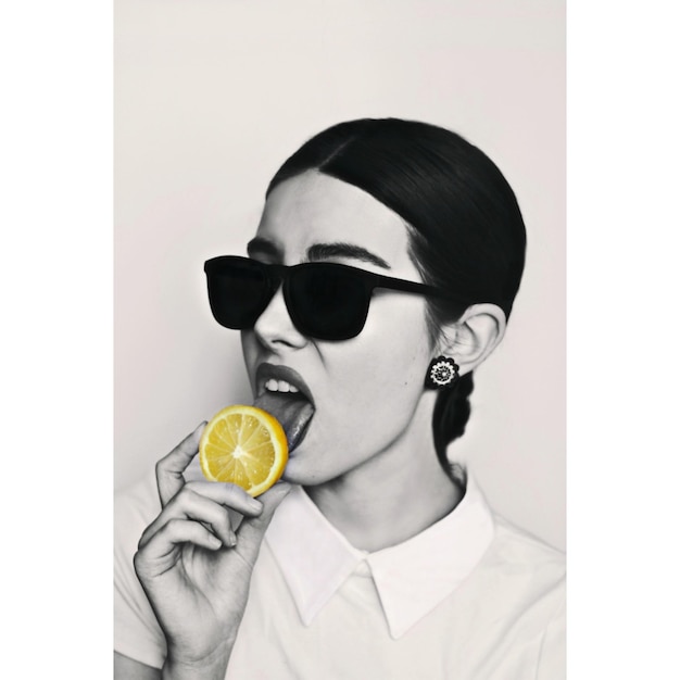Close-up of young woman wearing sunglasses while licking lemon