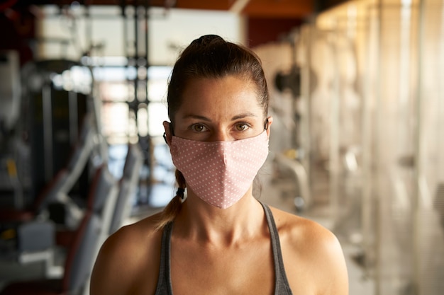 Photo close up of young woman with face mask looking at camera in gym.