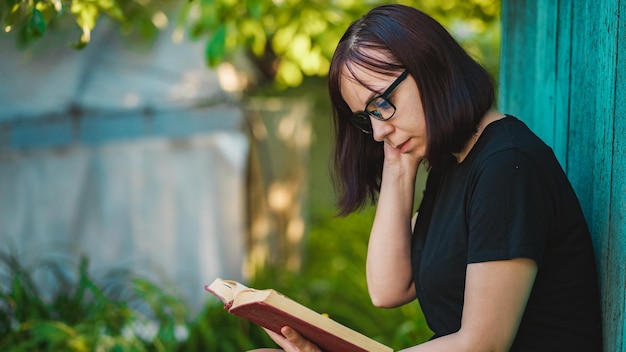 Close up of young woman with glasses reading book in garden Female resting in nature enjoying her leisure time