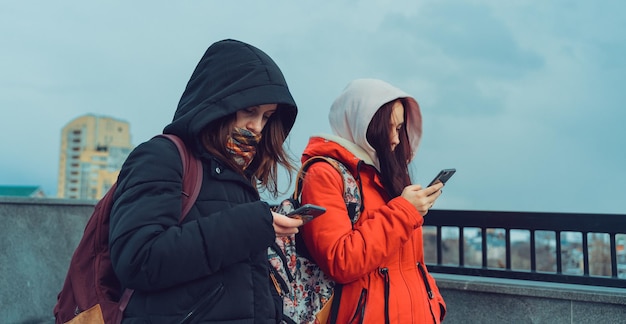 Close up of young women browsing smartphone in city park Pretty females in hoods using mobile phone