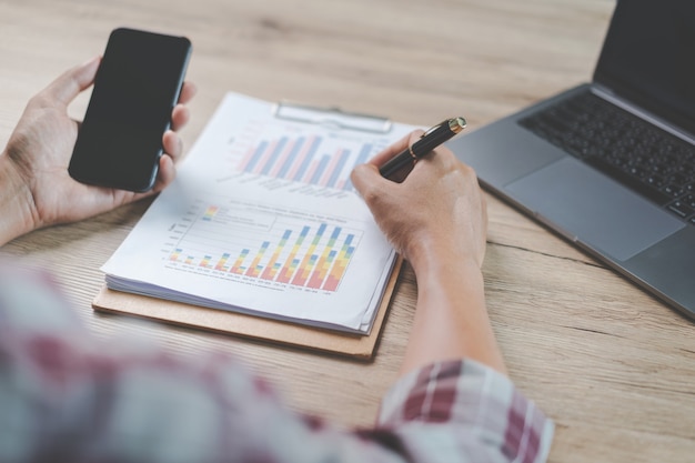 Close-up zakenman met een pen, smartphone en wijzend op financiële grafiek die bedrijfsrapport controleert op houten bureau met computerlaptop naast thuis. Stock foto