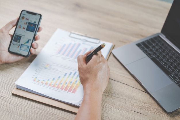 Close-up zakenman met een pen, smartphone en wijzend op financiële grafiek die bedrijfsrapport controleert op houten bureau met computerlaptop naast thuis. Stock foto