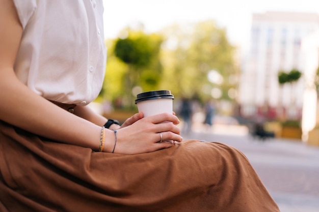 Close-up zijaanzicht bijgesneden schot van onherkenbare jonge vrouw die in handen papieren beker met afhaalkoffie zittend op een bankje op straat in de stad houdt