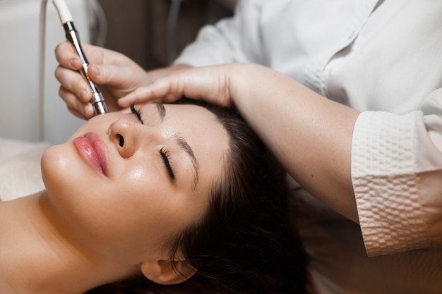 Close-up zijaanzicht portret van een leuke vrouw doet niet-invasieve microdermabrasie op haar gezicht met een dermapen door een schoonheidsspecialiste.