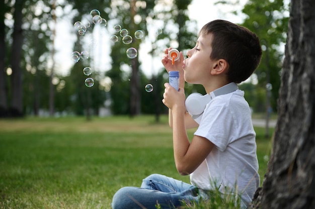 Close-up zijportret van een schattige vrolijke mooie schattige basisschooljongen die geniet van zijn recreatie tussen de lessen door, zeepbellen blazend, zittend op het groene gras van het stadspark