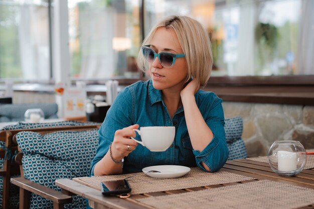 Close-up zomer portret van een jonge dame in modieuze zonnebril met een ochtend kopje koffie, heldere blauwe stijlvolle jurk.