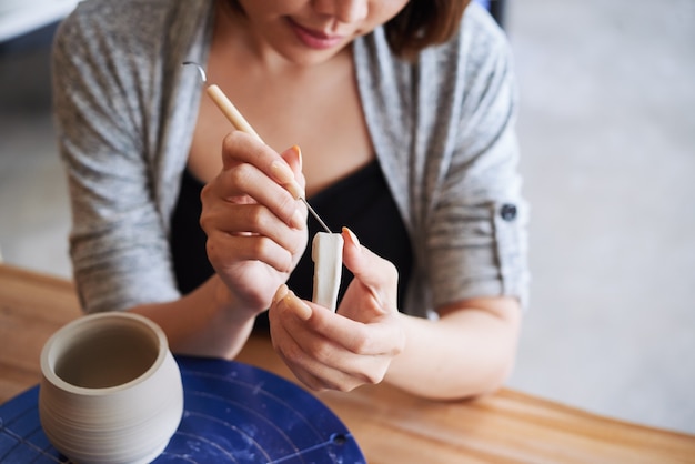 Close-upbeeld van creatieve jonge vrouw die handvat van kleikruik snijdt die zij op pottenbakkerswiel heeft gemaakt