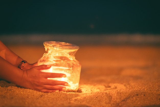 Close-upbeeld van de handen die van een vrouw en de houders van een glasfleskaarsen op het strand 's nachts houden