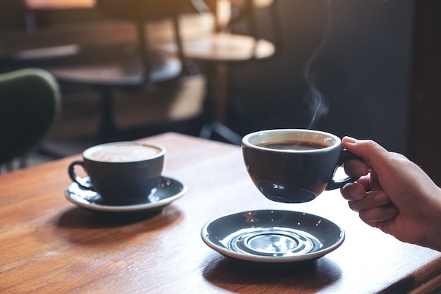 Close-upbeeld van een hand die een blauwe kop van hete koffie met rook op houten lijst in koffie houden