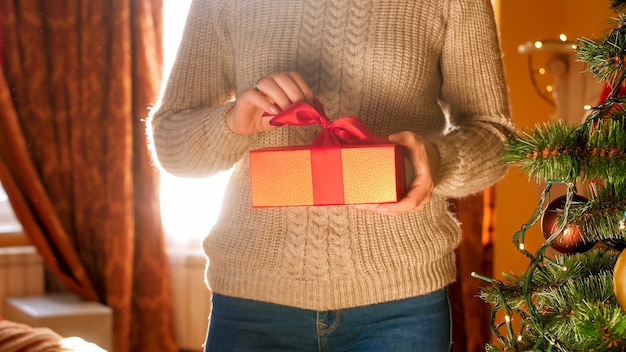 Close-upbeeld van een jonge vrouw die een doos met kerstcadeau vasthoudt en een strik trekt om het geschenk te openen