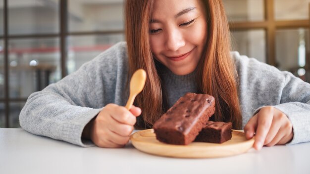 Close-upbeeld van een mooie aziatische vrouw die heerlijke browniecake in houten plaat kijkt en eet