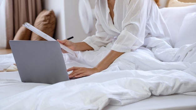 Close-upbeeld van een vrouw die een laptopcomputer gebruikt en aan papierwerk werkt terwijl ze thuis op bed zit