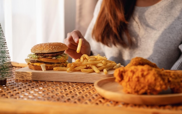 Close-upbeeld van een vrouw die frietjes en hamburger met gebakken kip op tafel thuis vasthoudt en eet