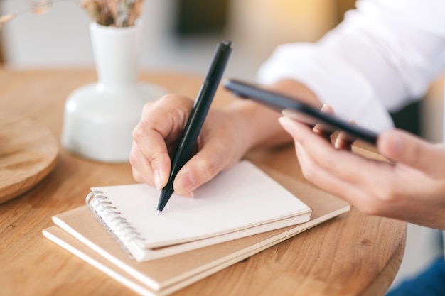 Close-upbeeld van een vrouw die mobiele telefoon gebruikt terwijl ze op een blanco notitieboekje op houten tafel schrijft