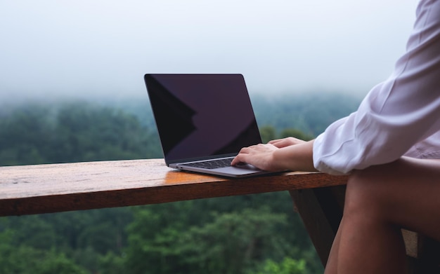 Close-upbeeld van een vrouw die op een mistige dag een laptop gebruikt en eraan werkt terwijl ze op een balkon zit met een prachtig uitzicht op de natuur