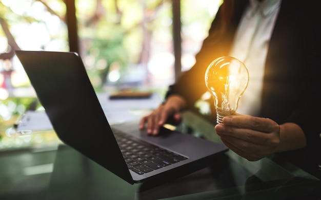 Close-upbeeld van een zakenvrouw die een gloeiende gloeilamp vasthoudt terwijl ze op een laptop op kantoor werkt