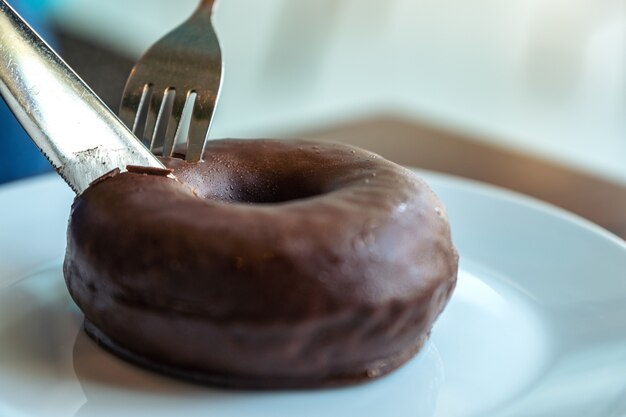 Close-upbeeld van handen die een stuk van chocoladedoughnut snijden door mes en vork voor ontbijt op houten lijst