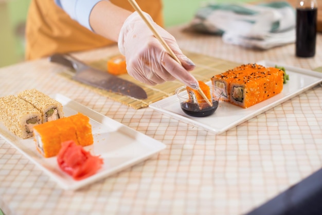Close-upbeeld van vrouwelijke hand in handschoen die een sushibroodje in eetstokjes houdt die het in sojasaus in keuken dompelen