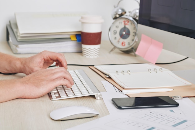 Close-upbeeld van zakenman die aan computer aan zijn bureau met geopende planner, smartphone en stapel dicuments werkt