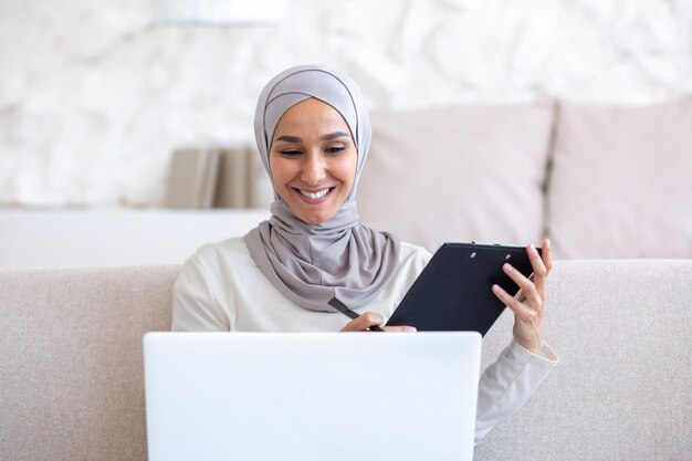 Close-upfoto van een jonge moslimvrouw in een hijab die thuis aan een laptop werkt, comfortabel op de bank zit en een map vasthoudt en op een videogesprek praat