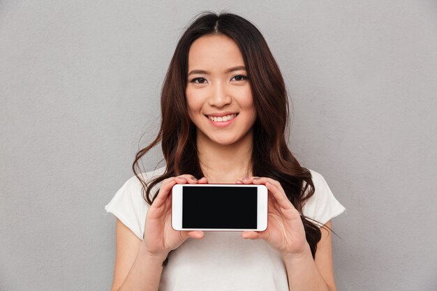 Close-upfoto van glimlachende Aziatische vrouw met lang bruin haar die het copyspacescherm van moderne smartphone tonen, dat over grijze muur wordt geïsoleerd