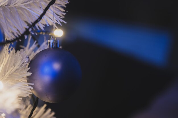 Close-upmening van blauwe vakantiebal die op witte kerstboom met glanzende lichten hangt