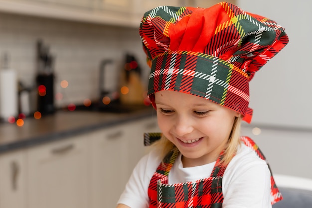 Close-upportret van een klein gelukkig glimlachend blondemeisje in een chef-kokkostuum in de keuken