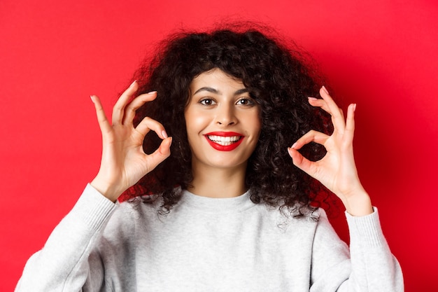 Close-upportret van glimlachende vrouw met krullend haar en rode lippen, die ok gebaar toont en tevreden kijkt, prijs goed product, die zich op rode achtergrond bevindt.